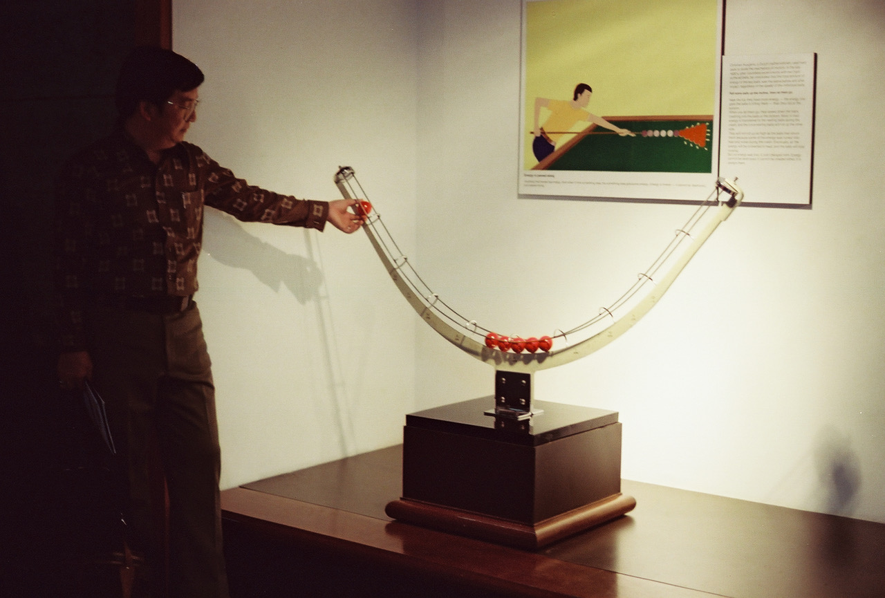 Dr. Chia releases a billiard ball, which will transmit its energy to the last ball on the opposite side, causing it to roll up the track.